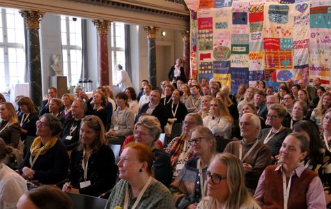 En stor gruppe mennesker sidder i et auditorium eller konferencerum med udsmykkede søjler og højt til loftet. Der hænger et farverigt kunstværk af sammensyede bannere på væggen til højre.