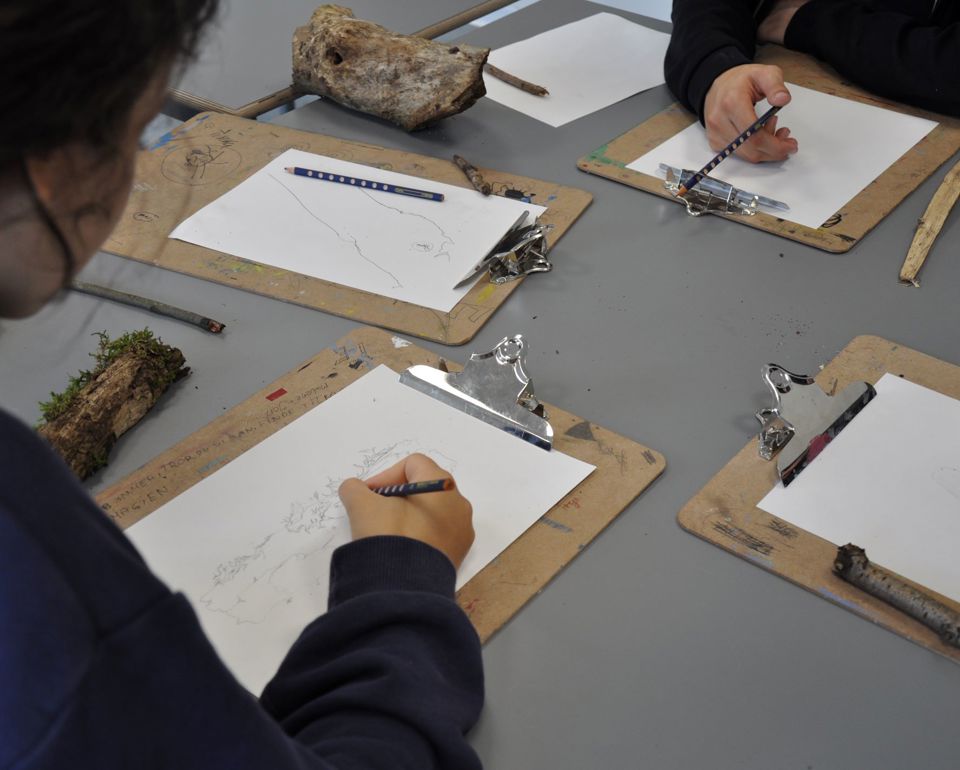 Flere børn sidder omkring et bord og tegner med blyanter på papir fastgjort til tegneplader med klemmer. På bordet ligger grene, bark og mos, som bruges som inspiration i en kreativ workshop.